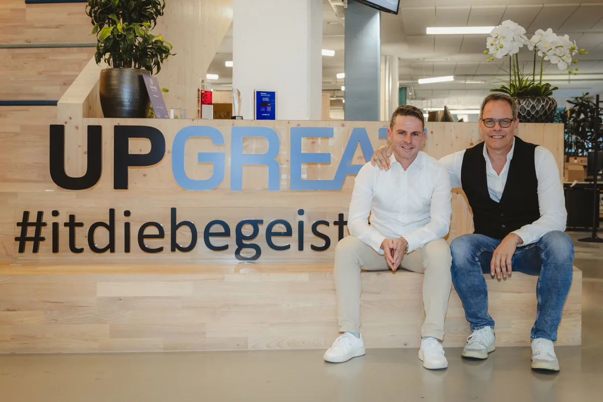 Simeon Roth (links) übernimmt bei Upgreat für Gründer Rinaldo Lieberherr. Zwei Männer sitzen vor einem hölzernen Korpus, der mit «Upgreat» beschriftet ist. Der Ältere legt seinen Arm auf die Schulter des Jüngeren.